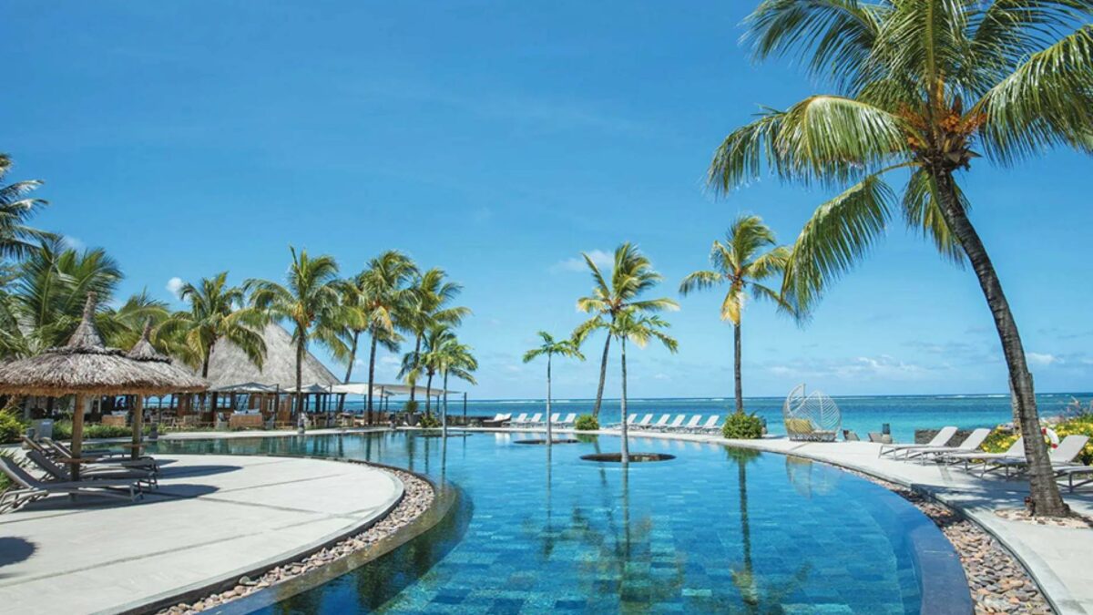 Luxurious beachfront resort in Mauritius featuring a stunning infinity pool surrounded by palm trees and thatched cabanas, overlooking the crystal-clear ocean under a bright blue sky.
