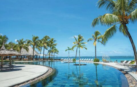 Luxurious beachfront resort in Mauritius featuring a stunning infinity pool surrounded by palm trees and thatched cabanas, overlooking the crystal-clear ocean under a bright blue sky.