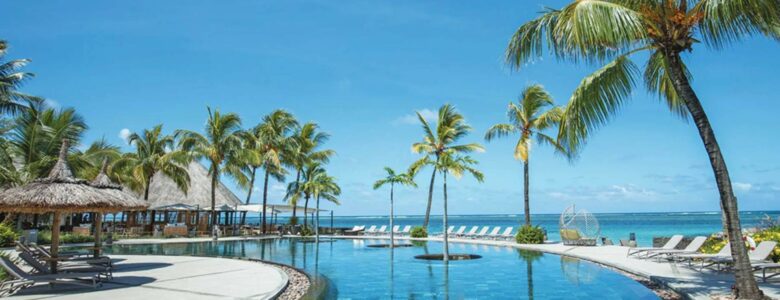 Luxurious beachfront resort in Mauritius featuring a stunning infinity pool surrounded by palm trees and thatched cabanas, overlooking the crystal-clear ocean under a bright blue sky.