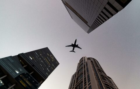 A view of an airplane flying overhead three skyscrapers. The view is long angled making the airplane seem framed in the middle of the skyscrapers. The sky above is grey. This blog is about Black Friday Flight Deals and Black Friday Travel Deals.