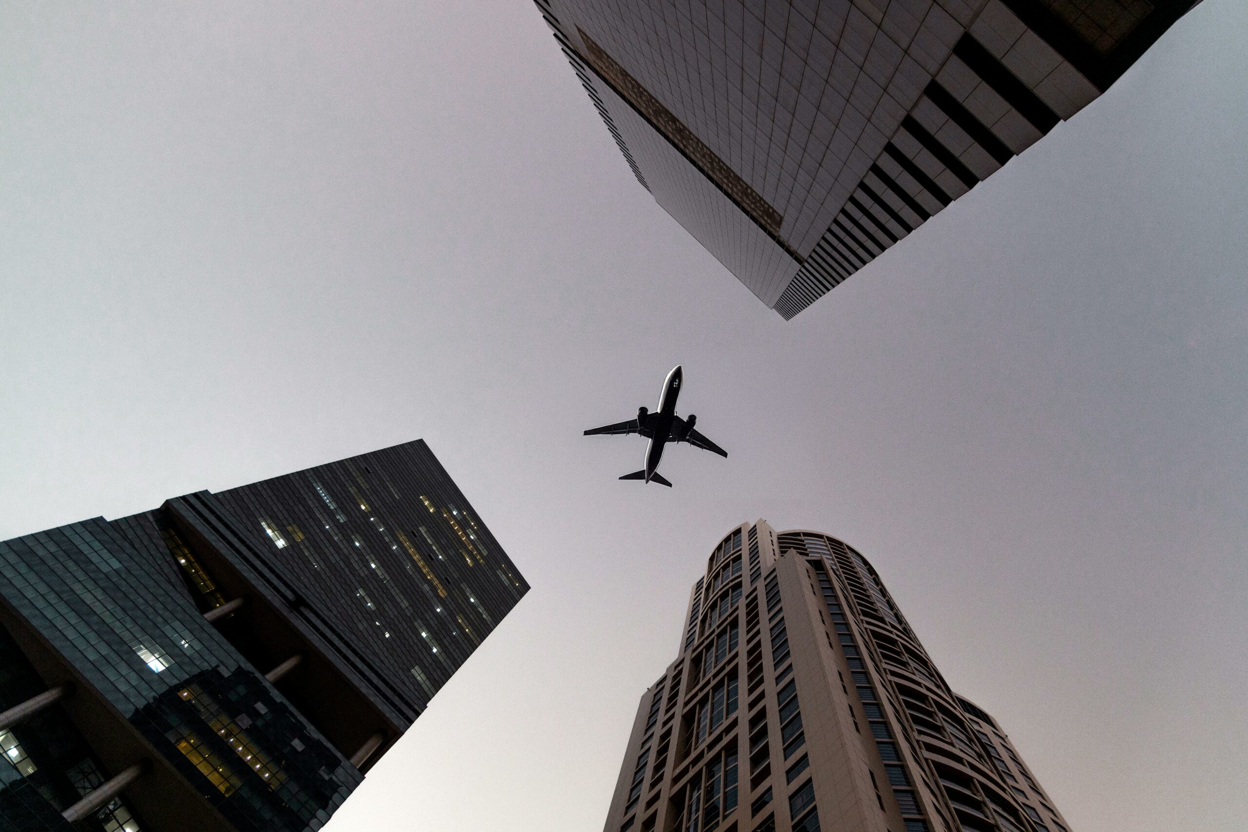 A view of an airplane flying overhead three skyscrapers. The view is long angled making the airplane seem framed in the middle of the skyscrapers. The sky above is grey. This blog is about Black Friday Flight Deals and Black Friday Travel Deals.