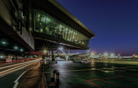 Night view showing a modern airport terminal with large glass windows at O.R Tambo International in Johannesburg. The runway is illuminated with bright lights, and several airplanes are parked nearby. The scene captures the bustling activity of an airport at night, highlighting the infrastructure and operations involved in air travel. This is for a blog titles the top 5 things to do during a layover in Johannesburg's O.R. Tambo International Airport