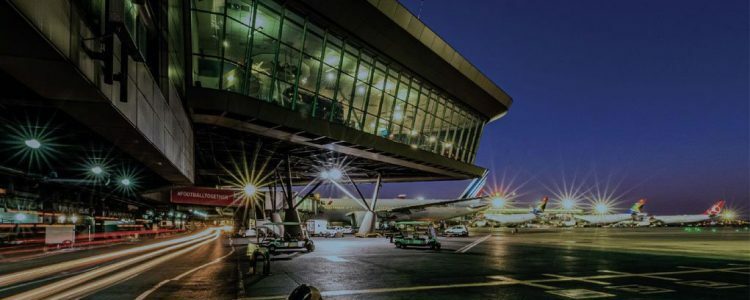 Night view showing a modern airport terminal with large glass windows at O.R Tambo International in Johannesburg. The runway is illuminated with bright lights, and several airplanes are parked nearby. The scene captures the bustling activity of an airport at night, highlighting the infrastructure and operations involved in air travel. This is for a blog titles the top 5 things to do during a layover in Johannesburg's O.R. Tambo International Airport