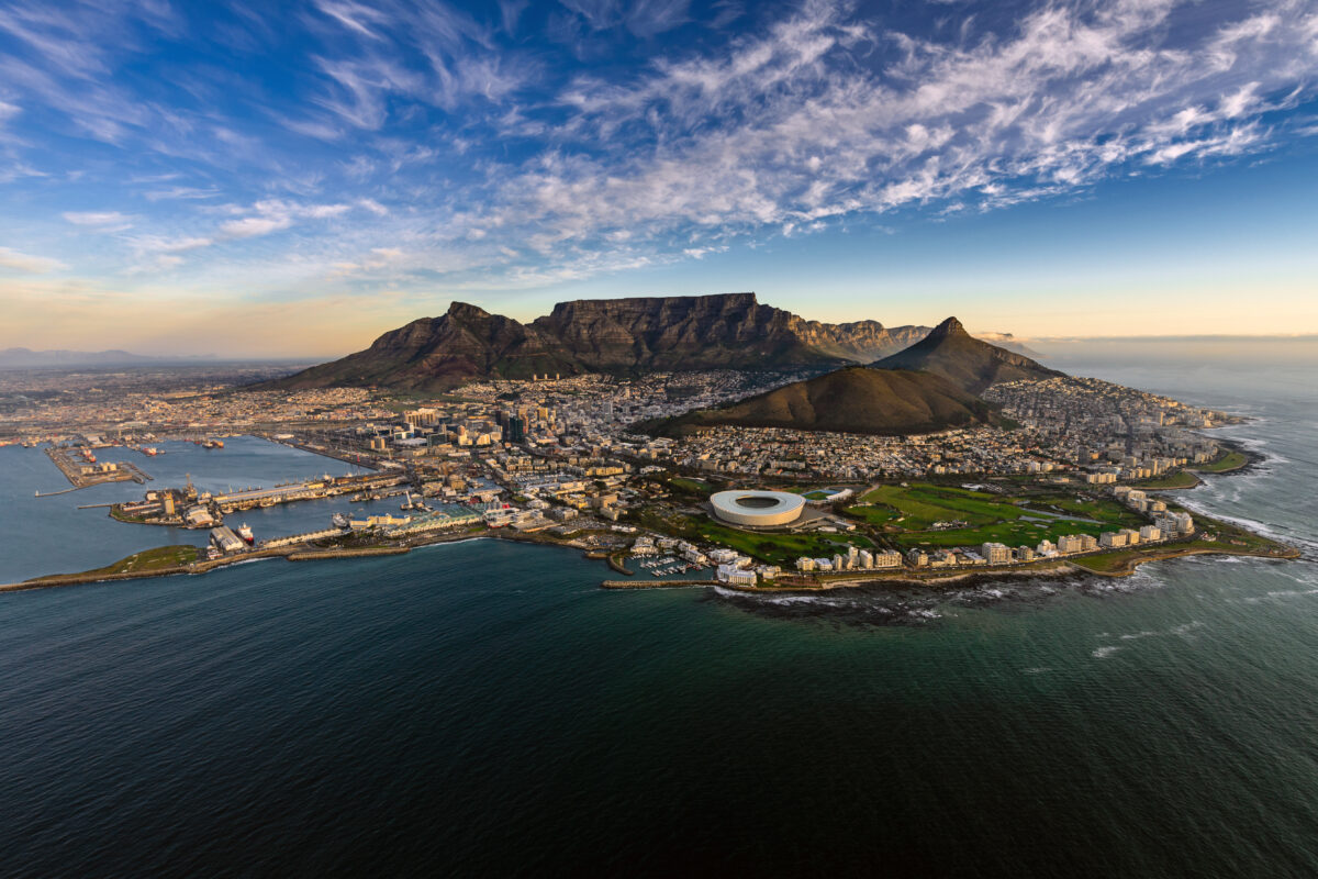 An Aerial view of Cape Town, voted the best city in the world 2025. In the foreground there is the ocean surrounding the city, in the middle is the city's architecture and stadium and in the background is the towering Table Mountain and Lion's Head an Chapman's Peak.
