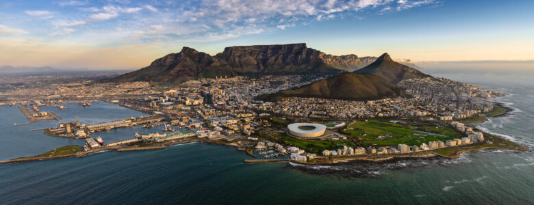 An Aerial view of Cape Town, voted the best city in the world 2025. In the foreground there is the ocean surrounding the city, in the middle is the city's architecture and stadium and in the background is the towering Table Mountain and Lion's Head an Chapman's Peak.