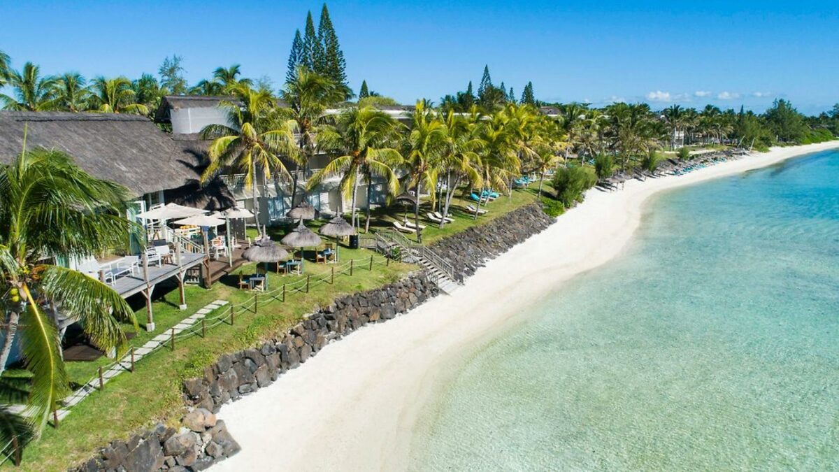 A stretched out section of holiday villas sheltered by tall palm trees in front of white beach sand and the blue ocean. These are places to see in Mauritius on a budget.