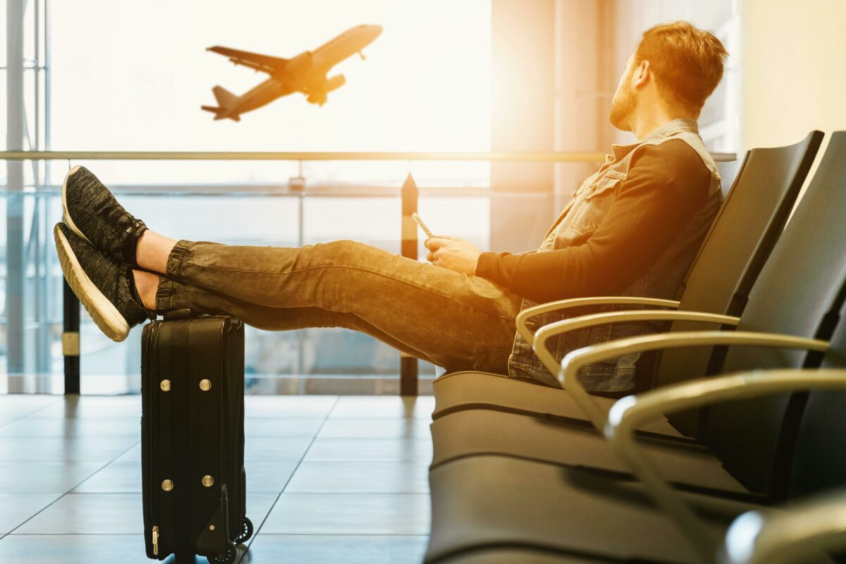 Man holding his phone while sitting in an airport looking at a plane taking off, probably googling "how to get a refund from an airline after a missed flight?"