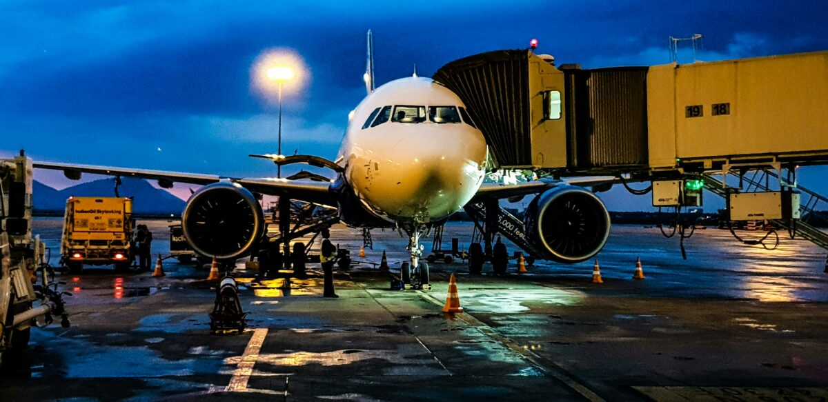 Commercial airplane at airport gate during evening boarding, with jet bridge connected and ground crew preparing for departure. This type of airport service is one of the reasons why flights are so expensive.