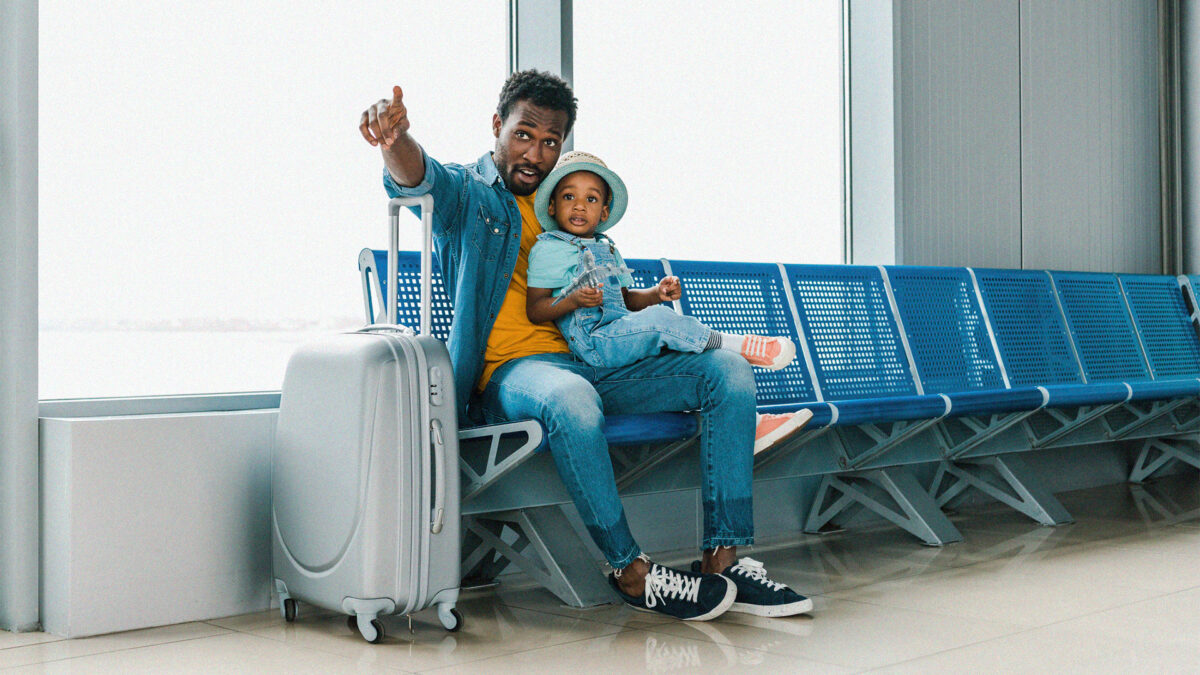 Father with his son at an airport, sitting on blue airport benches next to their suitcase. This is what's needed when one parent is travelling alone with the children.