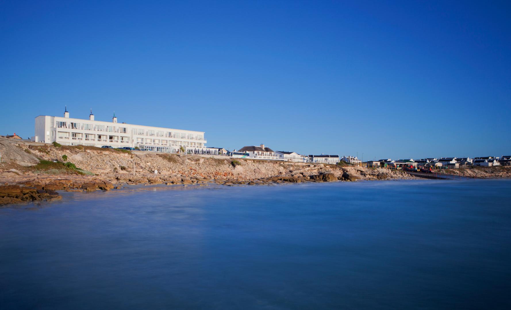 A coastal resort with a large white building, located on a rocky shoreline. The multi-story building features numerous windows and the surrounding area includes smaller houses extending along the coast under a clear blue sky and calm water, emphasizing the coastal landscape. This resort is the Arniston Spa Hotel in the Western Cape.