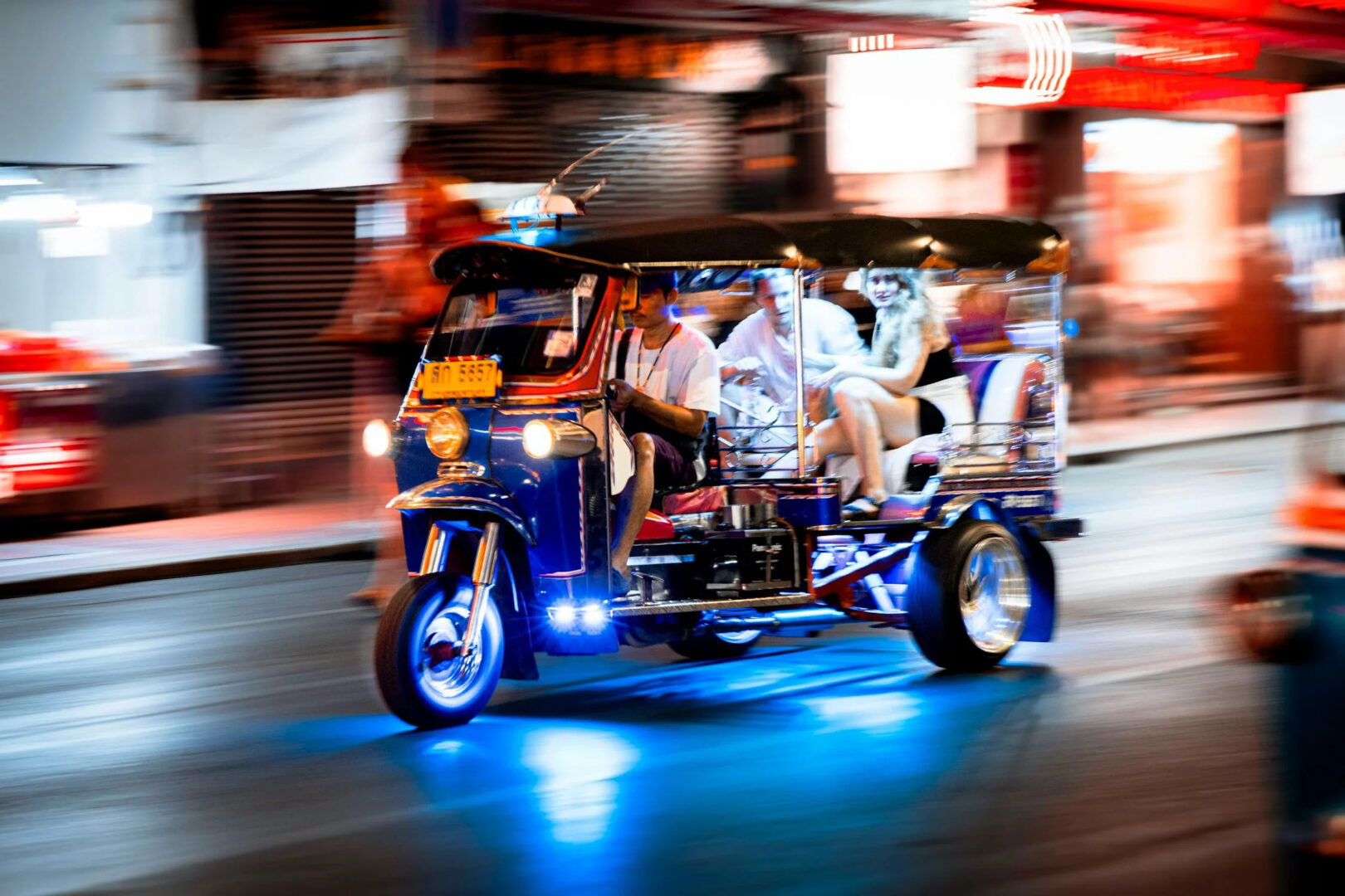 Bustling Bangkok nightlife on Khao San Road with neon lights. A tuk tuk carrying tourists zooming through the streets.