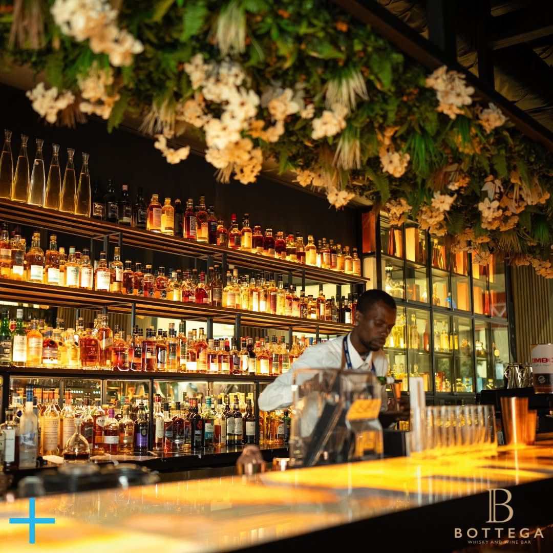 A bartender prepares drinks at Bottega Whisky and Wine Bar in Johannesburg's O.R. Tambo Airport. The bar is warmly lit and surrounded by shelves of premium liquor, with greenery hanging from the ceiling, creating a cozy and stylish atmosphere. This is one of the things to do during a layover in Johannesburg's O.R. Tambo Airport.