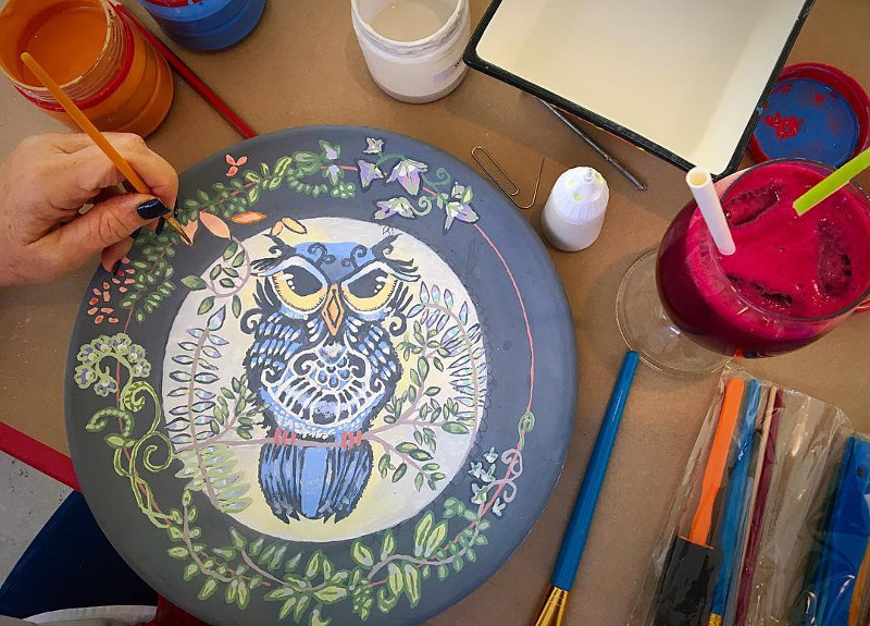 Person painting a colourful owl on a ceramic plate at a cosy Clay Café in Cape Town during winter, with paints scattered on the table.