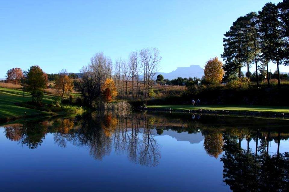 Champagne Sports Resort. A serene landscape featuring a calm lake reflecting the surrounding trees and clear blue sky. The scene includes a variety of trees with autumn foliage, a grassy area, and distant mountains. This location is one of the top 20 budget-friendly holiday resorts in South Africa for 2024 and 2025.