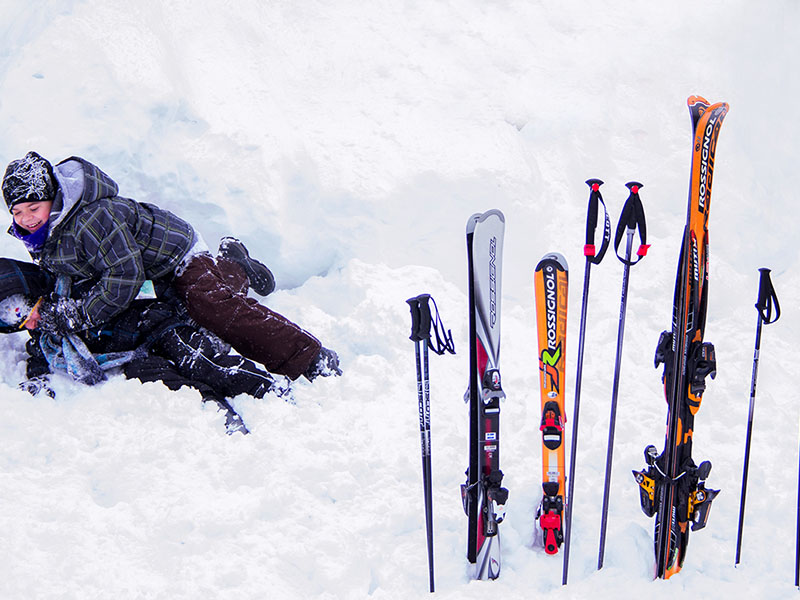 2 children dressed in winter wear playing in the snow and in the foreground there is skiing gear.