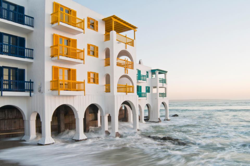 Club Mykonos in Langebaan, featuring white coastal buildings with colorful shutters and balconies in blue, yellow, and green. The buildings are built on arches extending into the water, with waves crashing against the base, showcasing the unique and vibrant architecture. This is one of the top 20 budget-friendly holiday resorts in South Africa for 2024 and 2025.