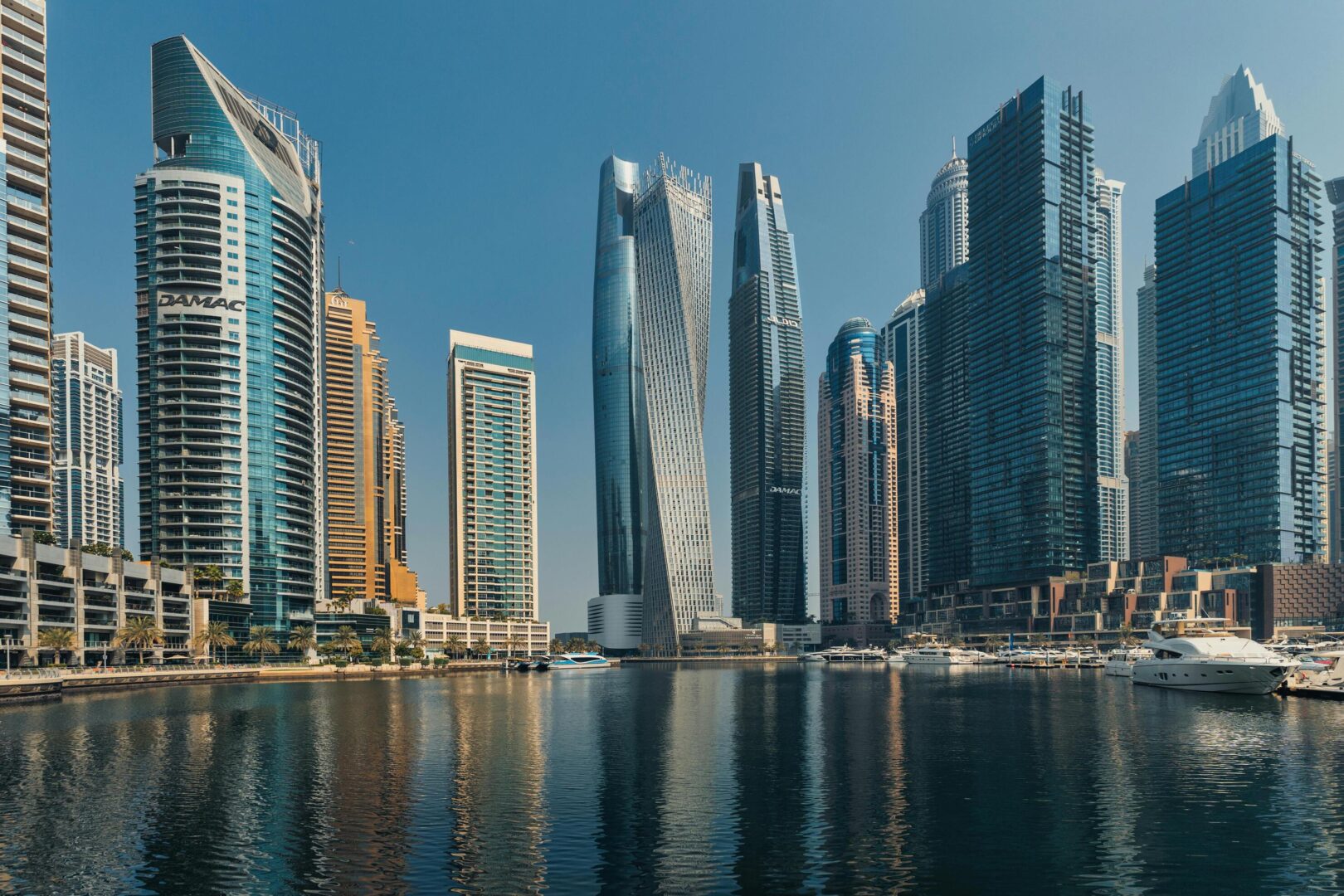 A daytime view of Dubai Marina with its iconic skyscrapers, including the twisting Cayan Tower, reflected in the calm waterfront. Luxury yachts line the marina, a hub for budget-friendly Dubai activities like strolling along the promenade, skyline photography, or enjoying the vibrant atmosphere. This is one of the free things to do in Dubai.