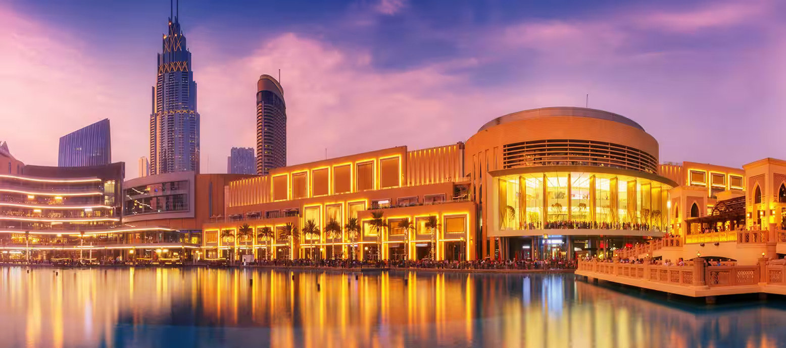 The Dubai Mall at sunset, illuminated by warm yellow lights that reflect on the still waters of the adjacent lake. The building's modern architecture features large glass windows and ornate details. This one of the most budget-friendly and free things to do in Dubai
