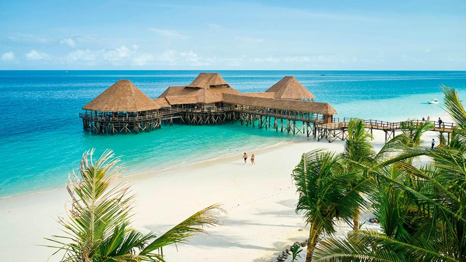 Brown thatched over-water huts on the blue sea next to white beach sand in Zanzibar, one of the affordable Easter holiday destinations for 2025