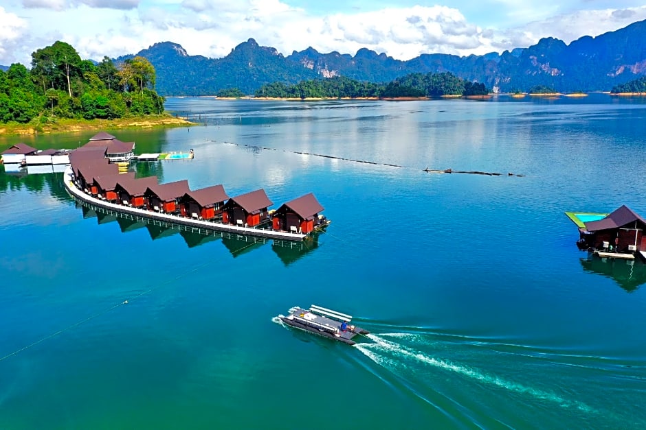 Floating villa huts in Thailand. A line of connected red huts on the blue sea with a tourist sailing near by. There are lush green mountains in the background.