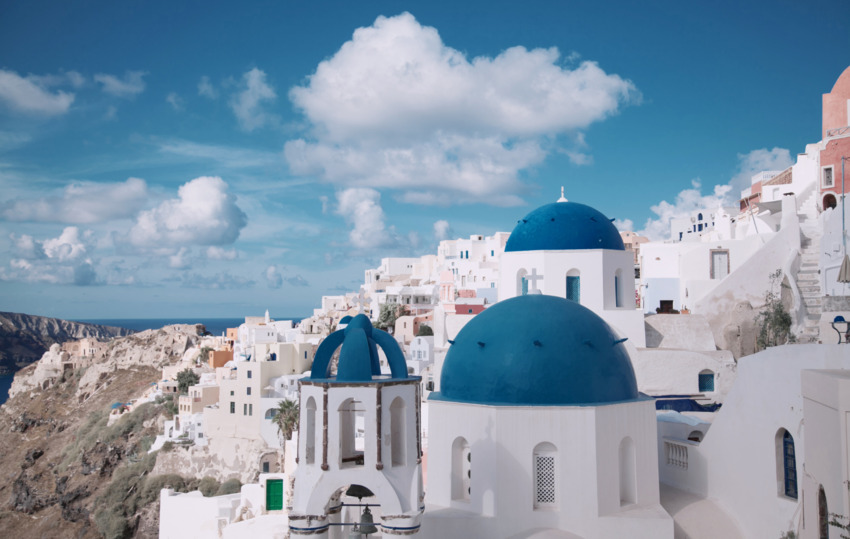 Scenic view of Santorini, Greece, there are white buildings with blue domed roofs under a bright blue sky and fluffy clouds. There is a mountain slope to the left side and a small view of the blue sea in the background. This blog is about Black Friday Flight Deals and Black Friday 2025 Travel Deals.