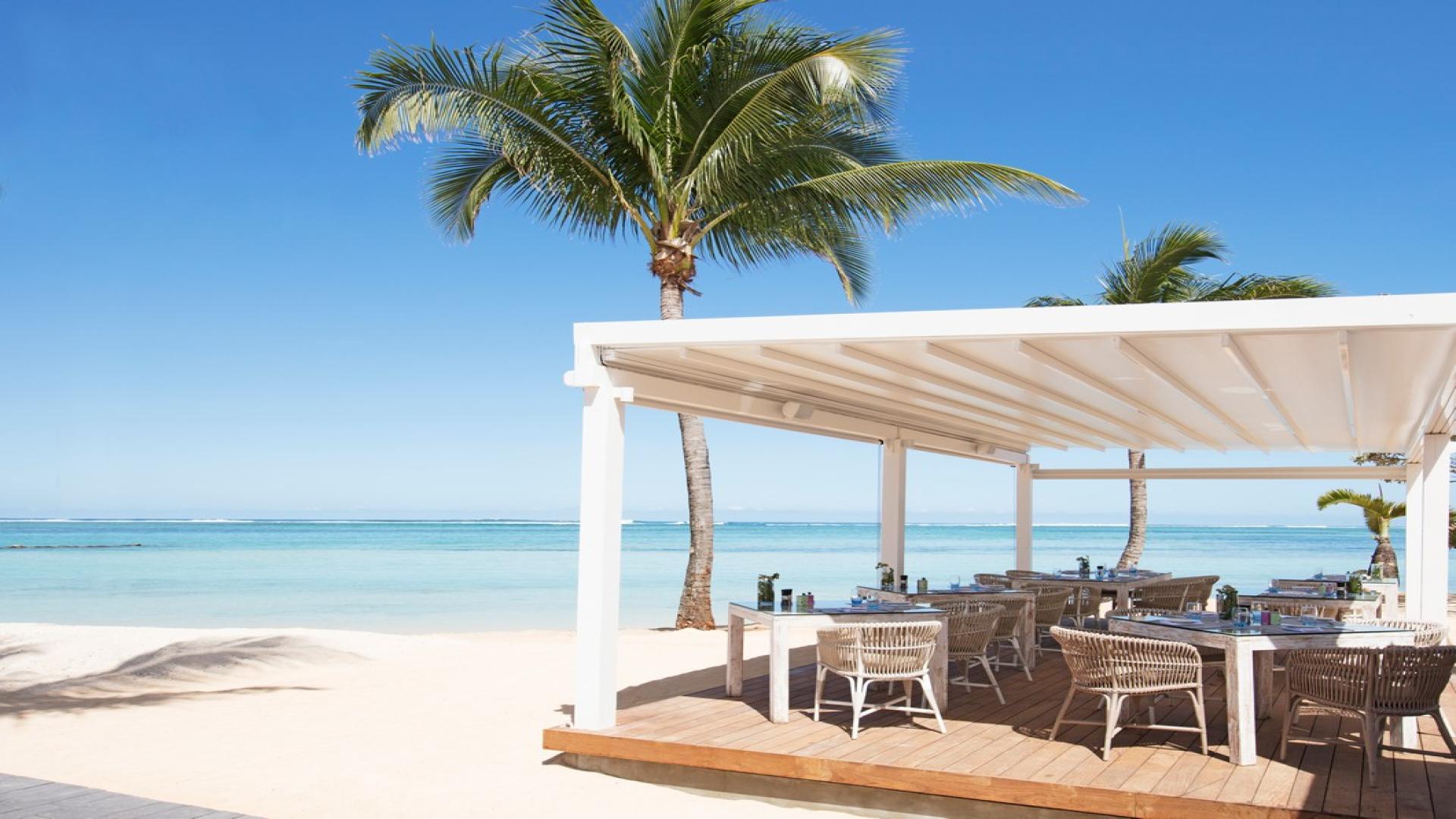 A white cabana sheltering a sitting area on white beach sand with tall palm trees in front of the crystal blue ocean and sky. These are places you'll see travelling to Mauritius on a budget.