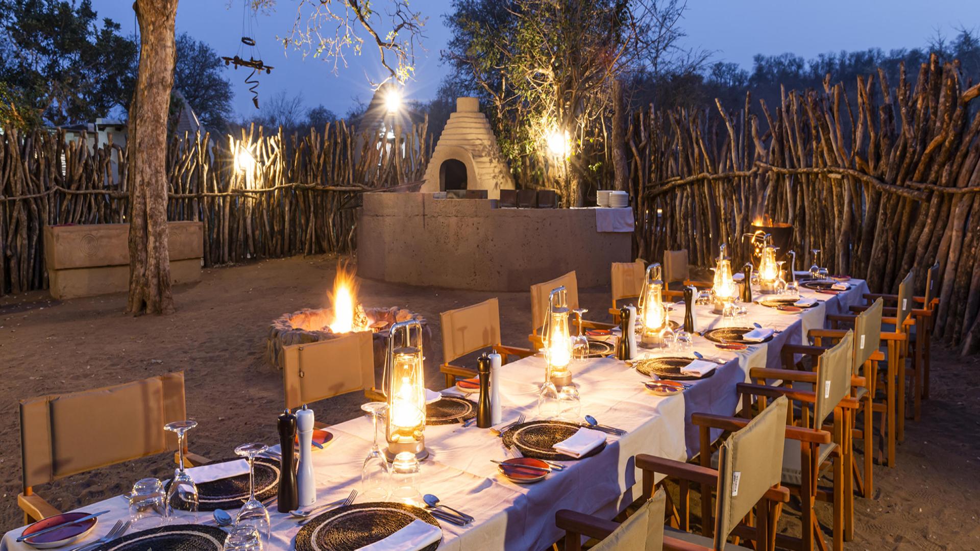 A rustic, outdoor dining area at dusk, set in a fenced African bush setting. There is a long table beautifully arranged with plates, cutlery, and lanterns, creating an inviting atmosphere. In the middle of the area there is a cozy fire pit. In the background there is a traditional African fence made with logs and tree bark. This resort is the Makalali Main Lodge in Limpopo and it is one of South Africa’s best Budget-Friendly Holiday Resorts for 2024 & 2025.