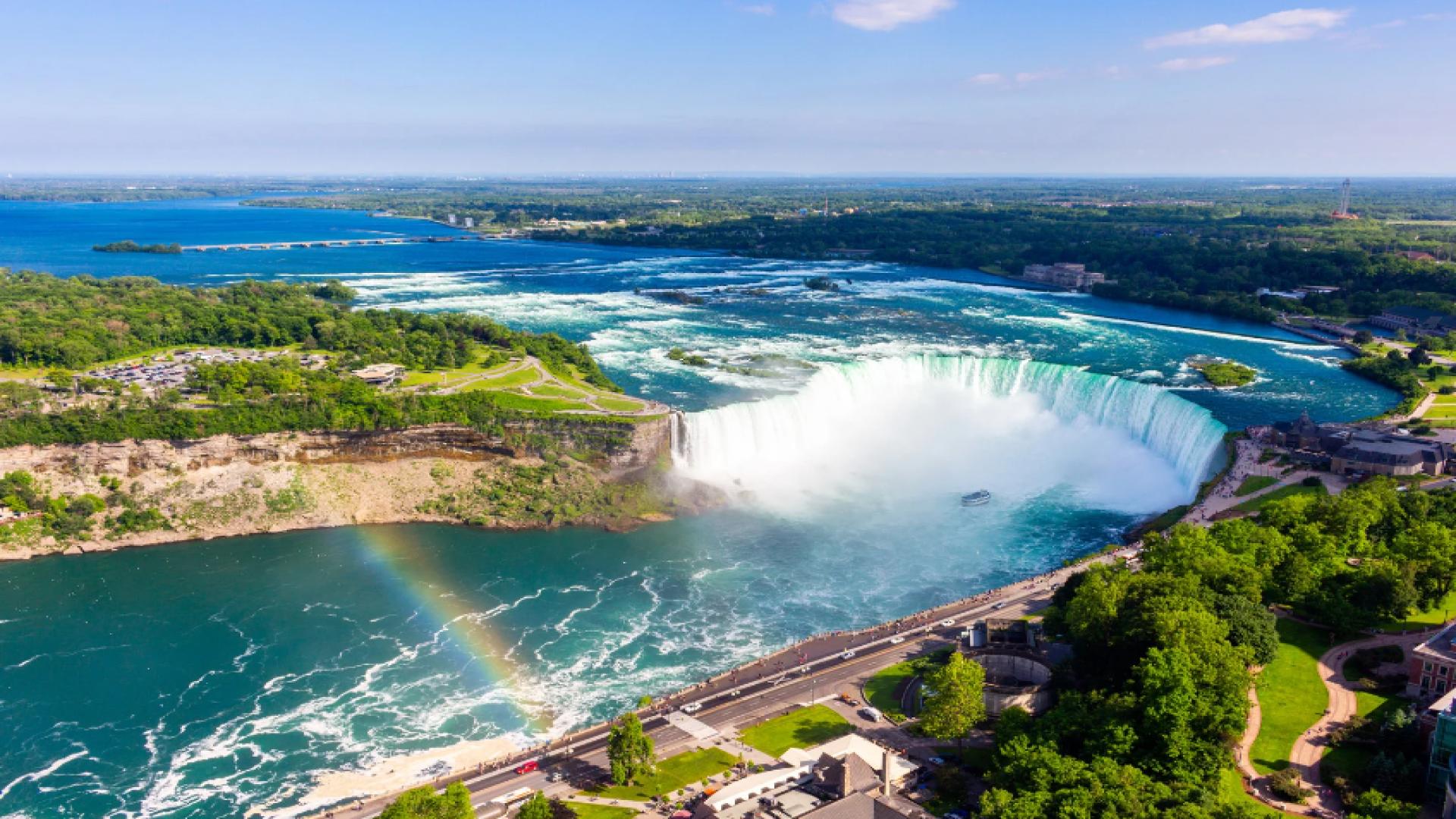 Aerial view of the Niagara Falls, featuring the cascading white waterfall, vibrant emerald-green river, and lush greenery of the surrounding landscape. A rainbow arcs through the mist.