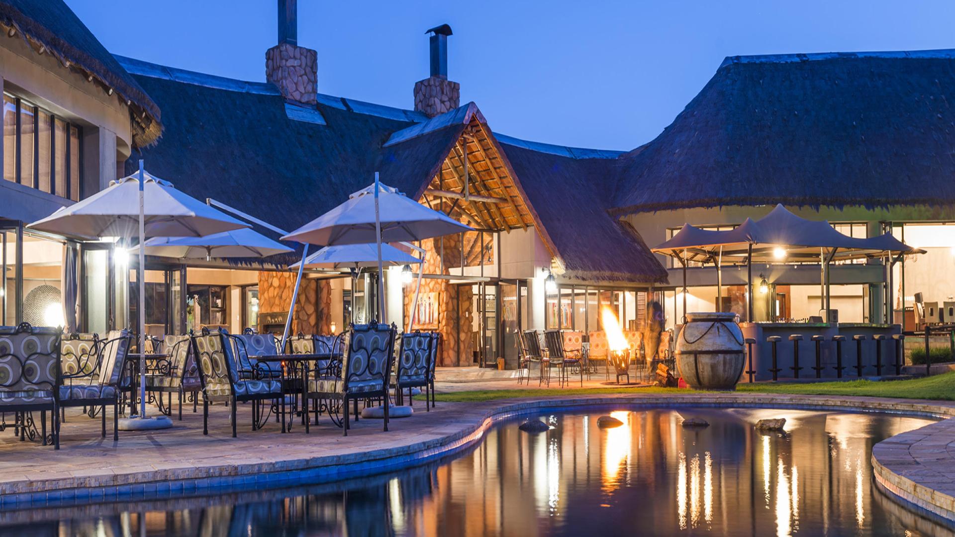 Luxurious resort at dusk featuring an elegant outdoor dining area with stylish chairs and tables under large umbrellas. The setting includes a thatched-roof building with expansive windows and stone accents, a cozy fire pit, and a tranquil swimming pool reflecting the ambient lights. This resort is the Ivory Tree Game Lodge in North West.
