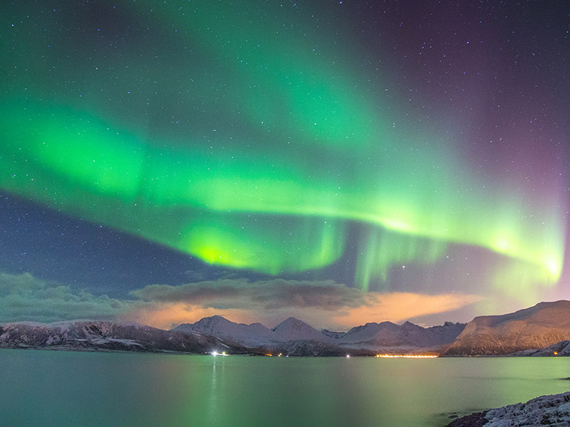 The northern lights at the Sommarøy Islands in Norway. The night sky is painted with vibrant green and purple hues and underneath the lights is mountainous landscape facing a large body of water which reflects the lights above.