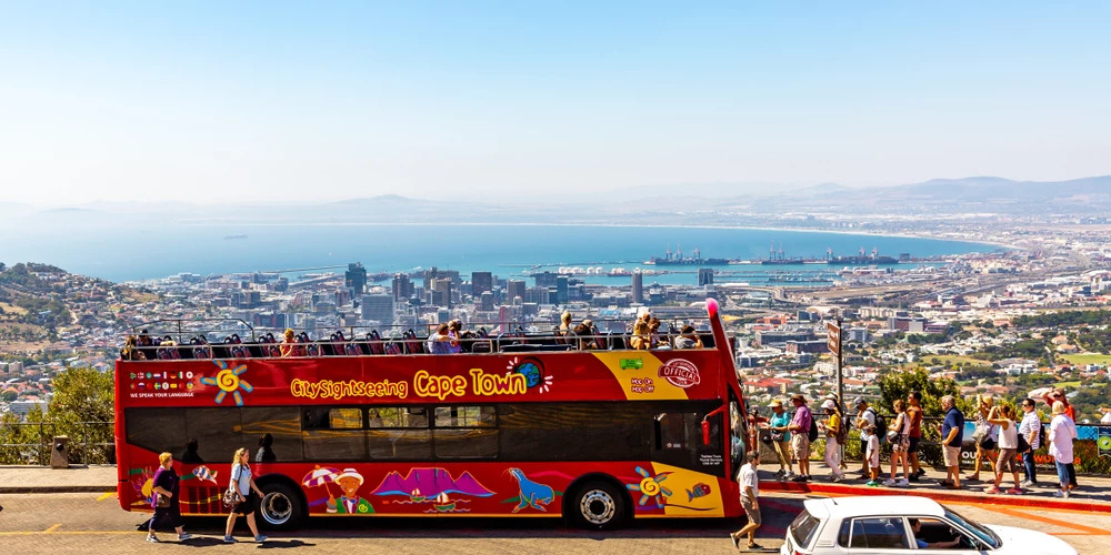 A red double-decker sightseeing bus, labeled 'City Sightseeing Cape Town,' is parked on a road with tourists onboard and nearby. The background showcases Cape Town's cityscape, coastline, and harbor. Taking a sightseeing bus tour is one of the cheap things to do in Cape Town.