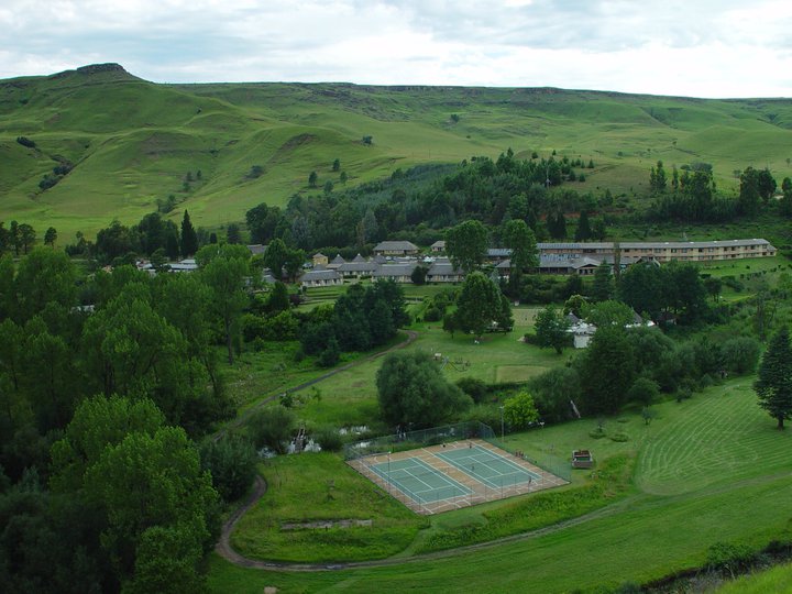 Scenic landscape featuring a well-maintained tennis court surrounded by lush green grass and trees. Behind the court, there is a large area with various trees and shrubs leading to a cluster of buildings. The background showcases rolling green hills stretching across the horizon. This serene setting is one of the top 20 budget-friendly holiday resorts in South Africa for 2024 & 2025. Sani Pass Hotel.