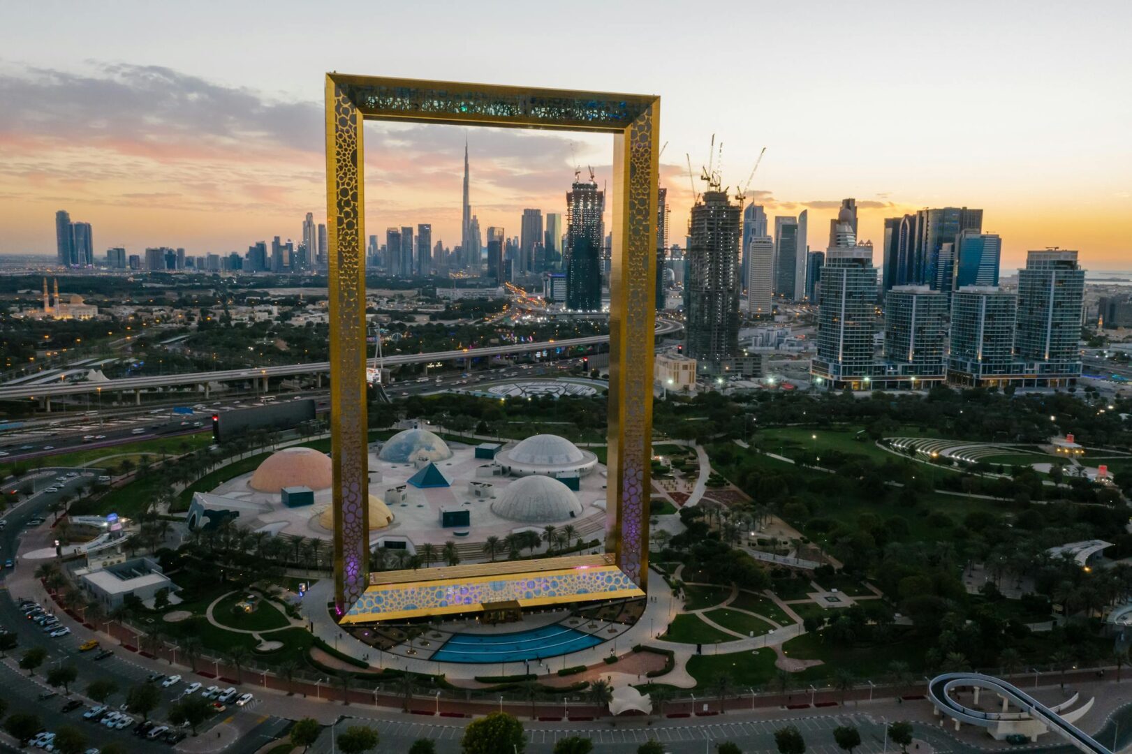 the grand Dubai Frame at sunset, a monumental golden rectangular structure with intricate patterns. In the background, the Dubai skyline is visible, featuring impressive skyscrapers including the iconic Burj Khalifa. The foreground showcases a park area with dome-shaped structures and pathways. The Dubai Frame, a significant architectural landmark, offers panoramic views of both old and new Dubai, making it an affordable and must-visit spot for those exploring budget-friendly Dubai.