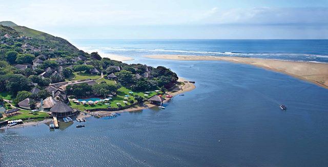 A scenic coastal area where the Umngazi river meets the ocean, surrounded by lush greenery. The area features numerous thatched-roof buildings which are part of the Umngazi resort, with well-maintained pathways and lawns. The calm water near the shore has a small boat visible. The coastline extends into the distance with sandy beaches and gentle waves. This is one of the top 20 budget-friendly holiday resorts in South Africa for 2024 and 2025.