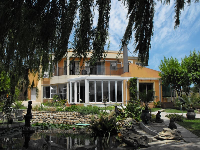 Two-story house with a yellow and white exterior, featuring a large glass-enclosed porch on the ground floor and a balcony on the upper floor. The house is surrounded by a well-maintained garden with various plants and trees, a stone pathway, and a pond with a statue in the foreground under a partly cloudy sky. This resort is the Valley of Peace in Stellenbosch.