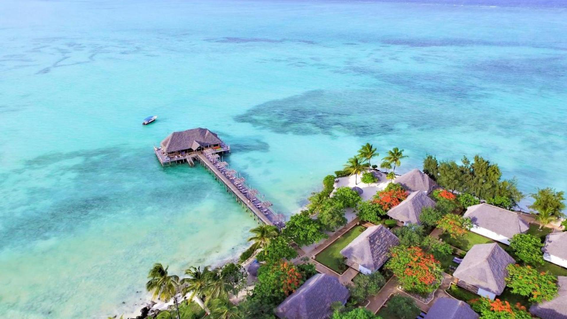 Aerial view of a tropical beachfront resort in Zanzibar, featuring turquoise blue ocean waters, lush palm trees, vibrant greenery, and overwater bungalows with thatched roof tops connected by a wooden pier. Plus one small boathouse on the ocean in the background. The picture has a very clam tone. Top 5 cheap Rand-frienldy travel deals for Black Friday 2025