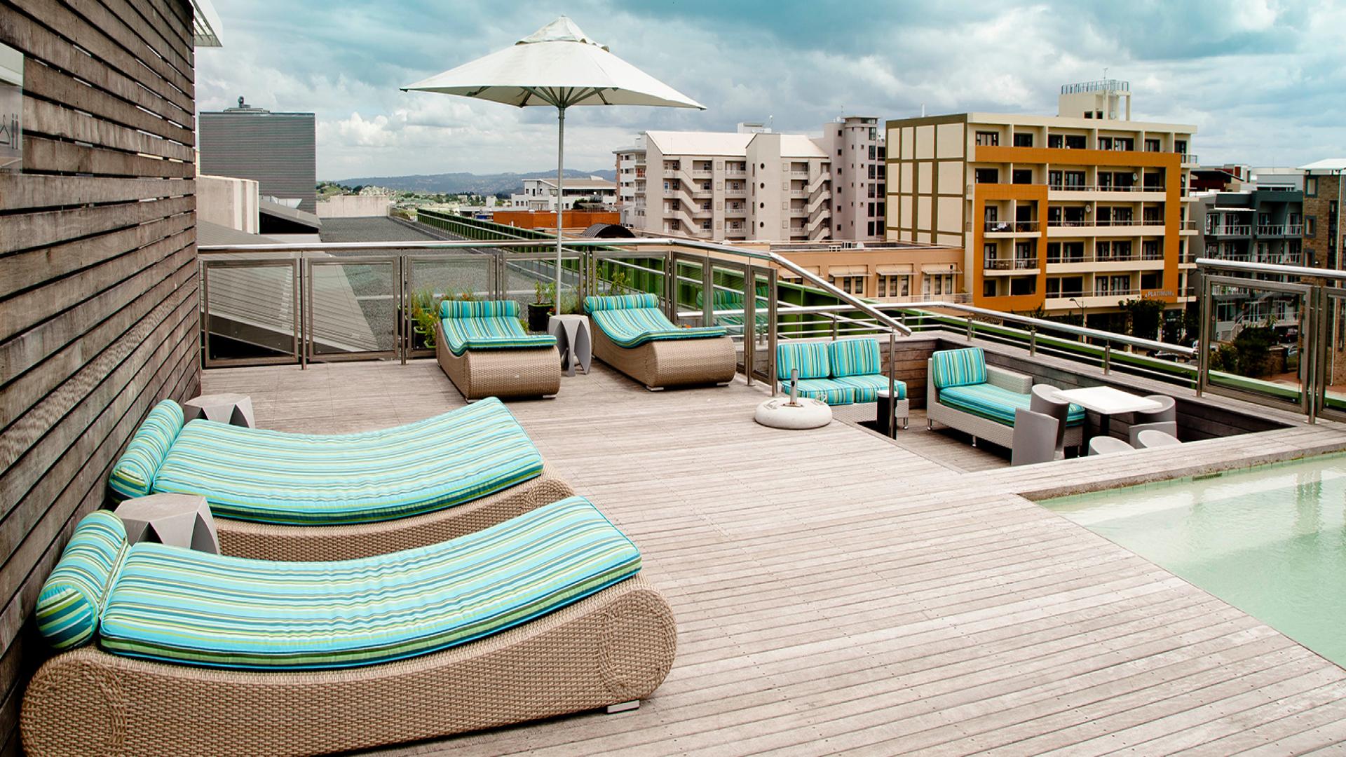 Modern rooftop terrace with wicker lounge chairs and turquoise striped cushions, a large umbrella, and a small pool, overlooking urban buildings. The terrace provides a scenic and relaxing outdoor space amid a bustling cityscape. This resort is the aha Gateway Hotel in KZN and it is one of South Africa’s best Holiday Resorts for 2024 & 2025.