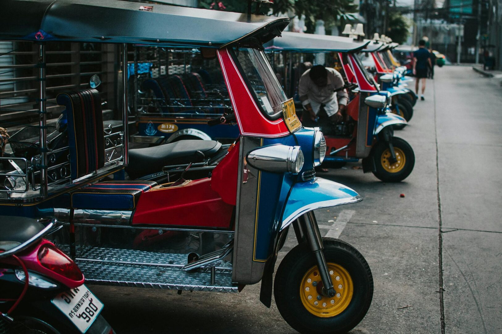A line of tuk tuk vehicles parked on a street in Phuket. They red framed with blue hints at the front and sliver flooring. Taking a tuk tuk is the cheapest mode of transportation for getting around Thailand on a budget.