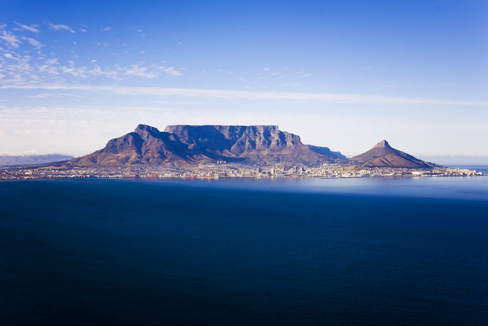 A panoramic view of Cape Town with Table Mountain and Lion's Head in the background. The cityscape includes buildings and roads at the base of the mountains, with the ocean in the foreground. The clear blue sky and expansive view of the ocean highlight the beauty of this iconic landscape. Visiting Table Mountain is one of the best things to do in Cape Town.