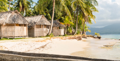 Sunny tropical beach with beach huts on piles with thatched roof for tourists and some coconut palm trees at the side. Canoe in front view. Panama